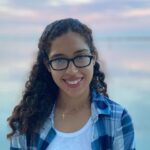 Crest Scholar Carolina Mahecha Millan smiles in front of a scenic, pale pink and powder blue sunset. She wears glasses, a white shirt, and blue flannel.