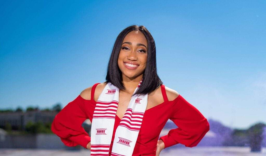 Crest Scholar Maya Jones smiles as she poses in front of a blue sky and a picturesque college campus. She's wearing a red dress.