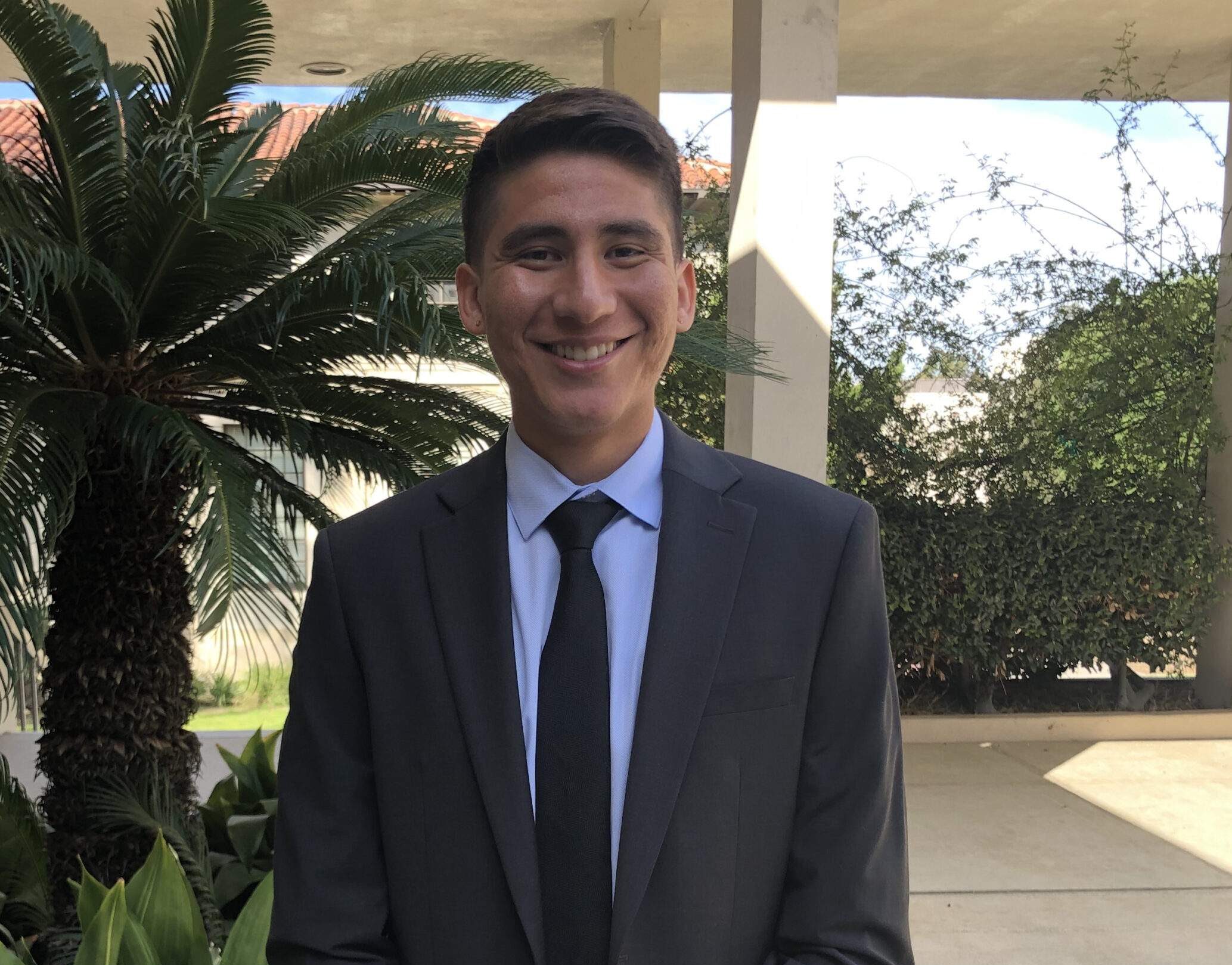 Crest scholar Oliver Artiga is wearing a suit and smiling while standing in front of a background of plants.