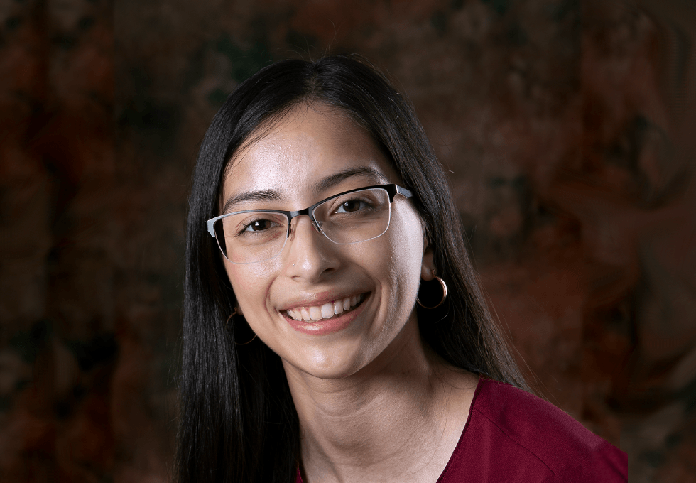 Crest Scholar Erika Gonzalez smiles for a professional portrait against a brown and black background. She wears glasses and a maroon shirt.
