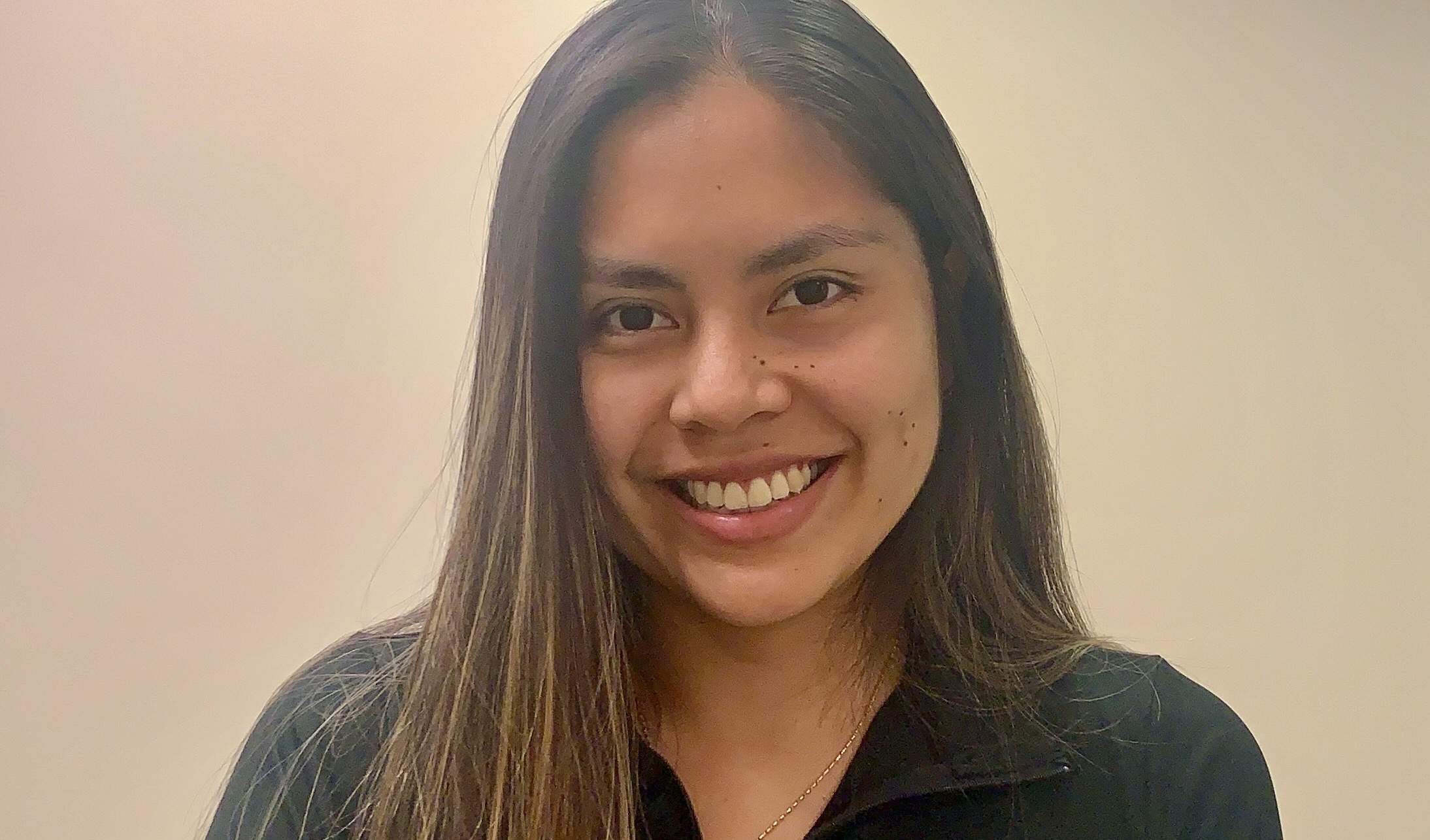 Crest scholar Fiorella Barreda smiles while standing in front of a plain wall. She's wearing a black, long-sleeved polo that says "Elon Doctor of Physical Therapy"