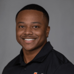 Keairez Coleman smiles for a professional headshot in front of a gray background. He's wearing a black polo shirt.