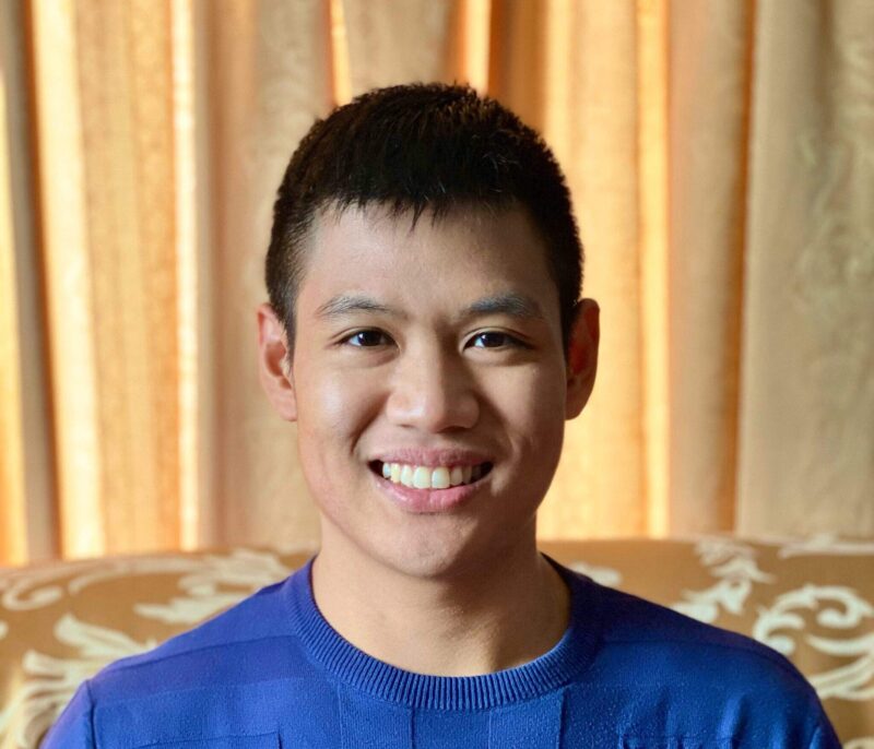 Crest Scholar Liam Calvin Henson smiles in front of a beige fabric background. He wears a blue t-shirt.