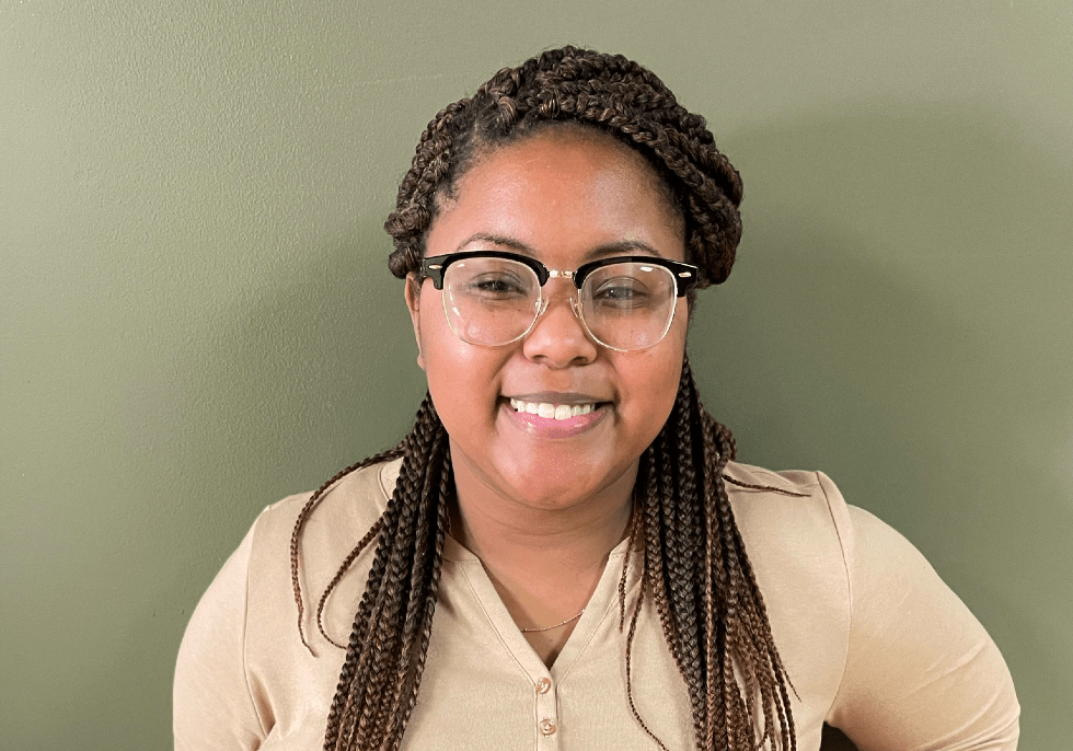 Crest Scholar Mikaela Holley smiles for a headshot while posing against a blank, beige wall. She's wearing glasses and a beige long-sleeved button-up.