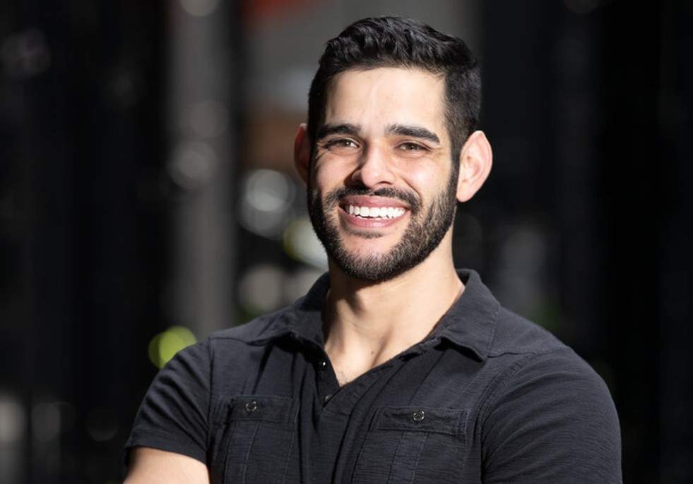 Ricki Locci smiles and poses with his arms crossed for a professional headshot. He's wearing a black short-sleeved polo.