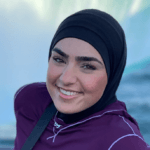 Crest Scholar Zahraa Darwich smiles at the camera while posing in front of scenic waterfalls. She's wearing a black hijab and a purple long-sleeve shirt