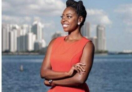 Crest scholar Brittany Ballentine smiles in front of a river and a city skyline. She's wearing an orange dress and her arms are crossed as she looks into the distance.