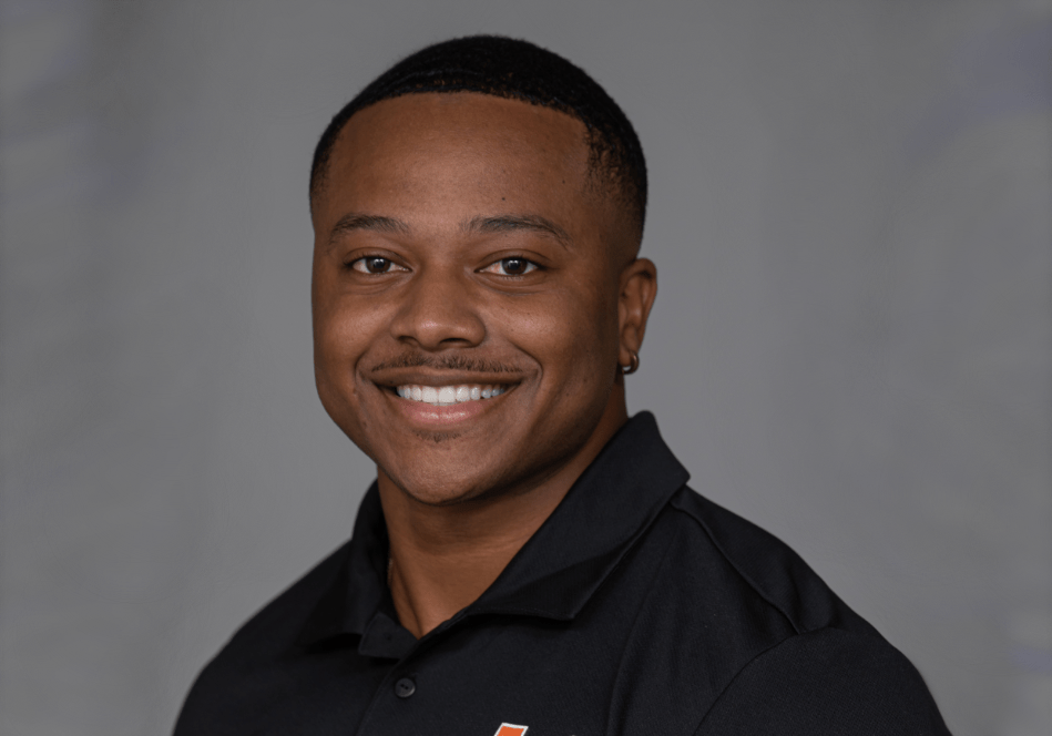 Keairez Coleman smiles for a professional headshot in front of a gray background. He's wearing a black polo shirt.