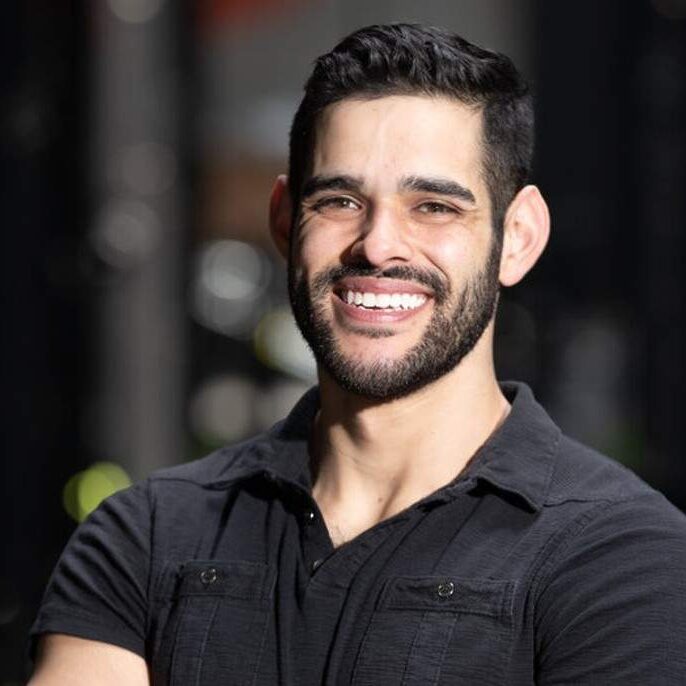 Ricki Locci smiles and poses with his arms crossed for a professional headshot. He's wearing a black short-sleeved polo.